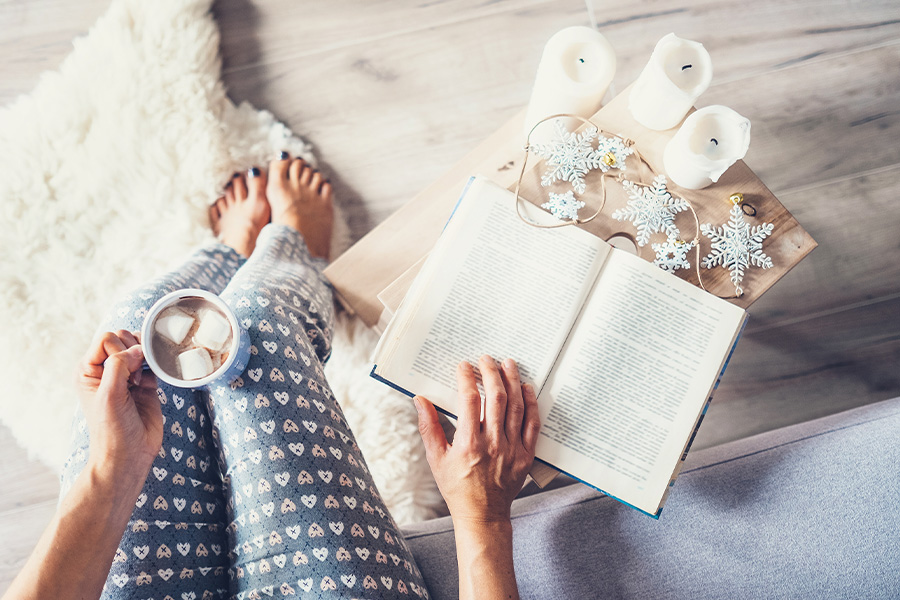 Kvinna läser en bok i soffan med julpynt och ljus på ett bord.